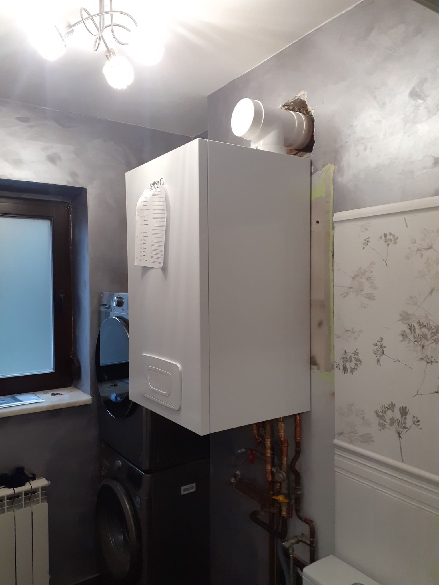 Biały piec gazowy zamontowany w łazience, widoczne rury przyłączeniowe, obok pralka, ściana z efektem betonu i fragment tapety w kwiaty.