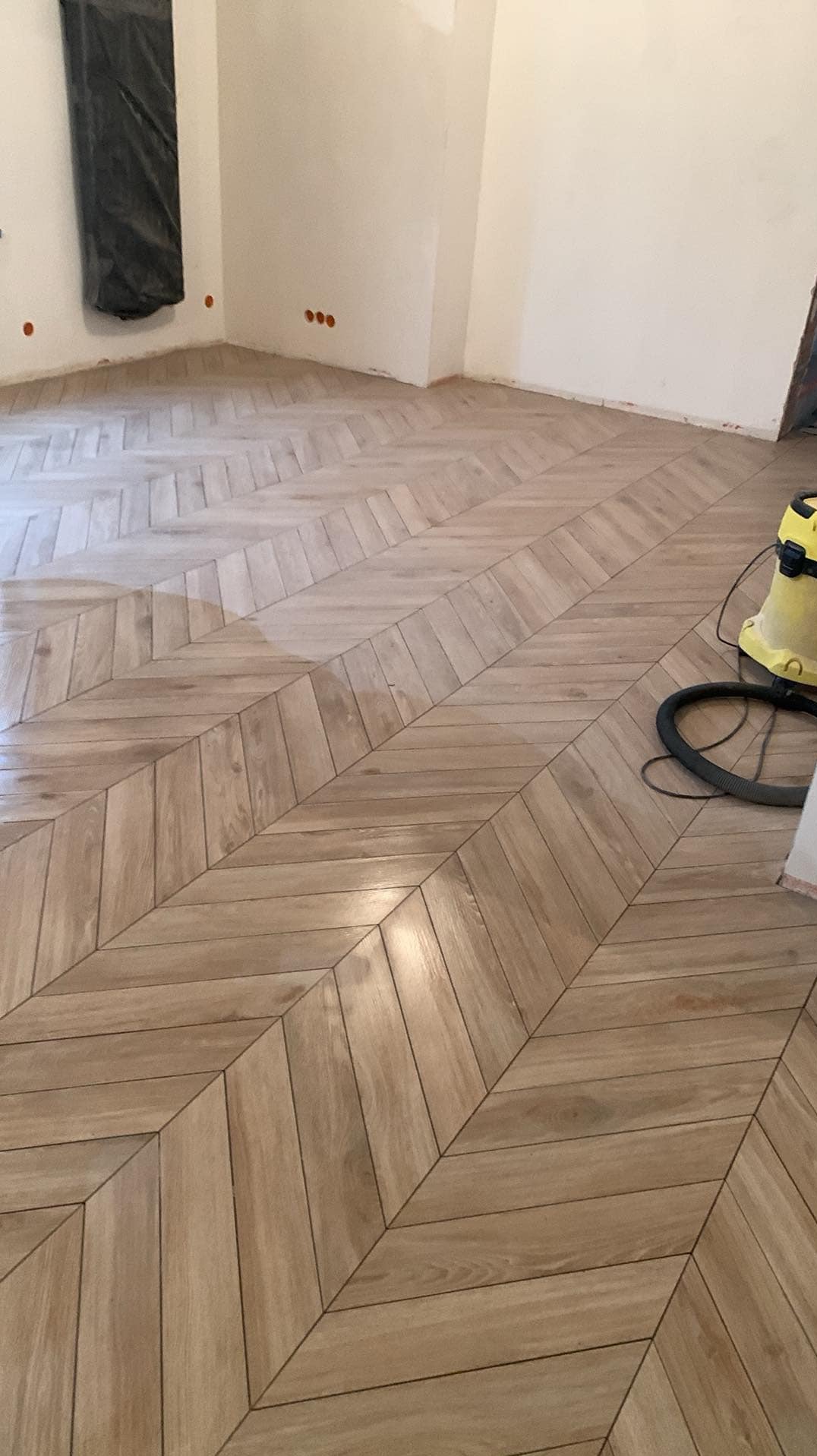 Ułożona w jodełkę jasna podłoga z paneli, na ścianie widoczne gniazdka elektryczne i czarny worek, obok żółty odkurzacz.