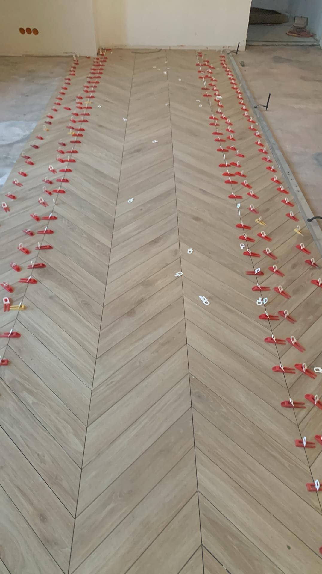 Ułożona pod kątem jasna podłoga z płytek imitujących drewno, z widocznymi czerwonymi i białymi klipsami poziomującymi płytki, wskazującymi na trwający proces montażu.