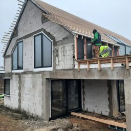 INVESTPROJEKT - Budowa dwukondygnacyjnego domu z białych bloczków, widoczne duże okna z czarnymi ramami, drewniana konstrukcja dachu w trakcie montażu, robotnicy pracujący na drewnianym rusztowaniu przy montażu...