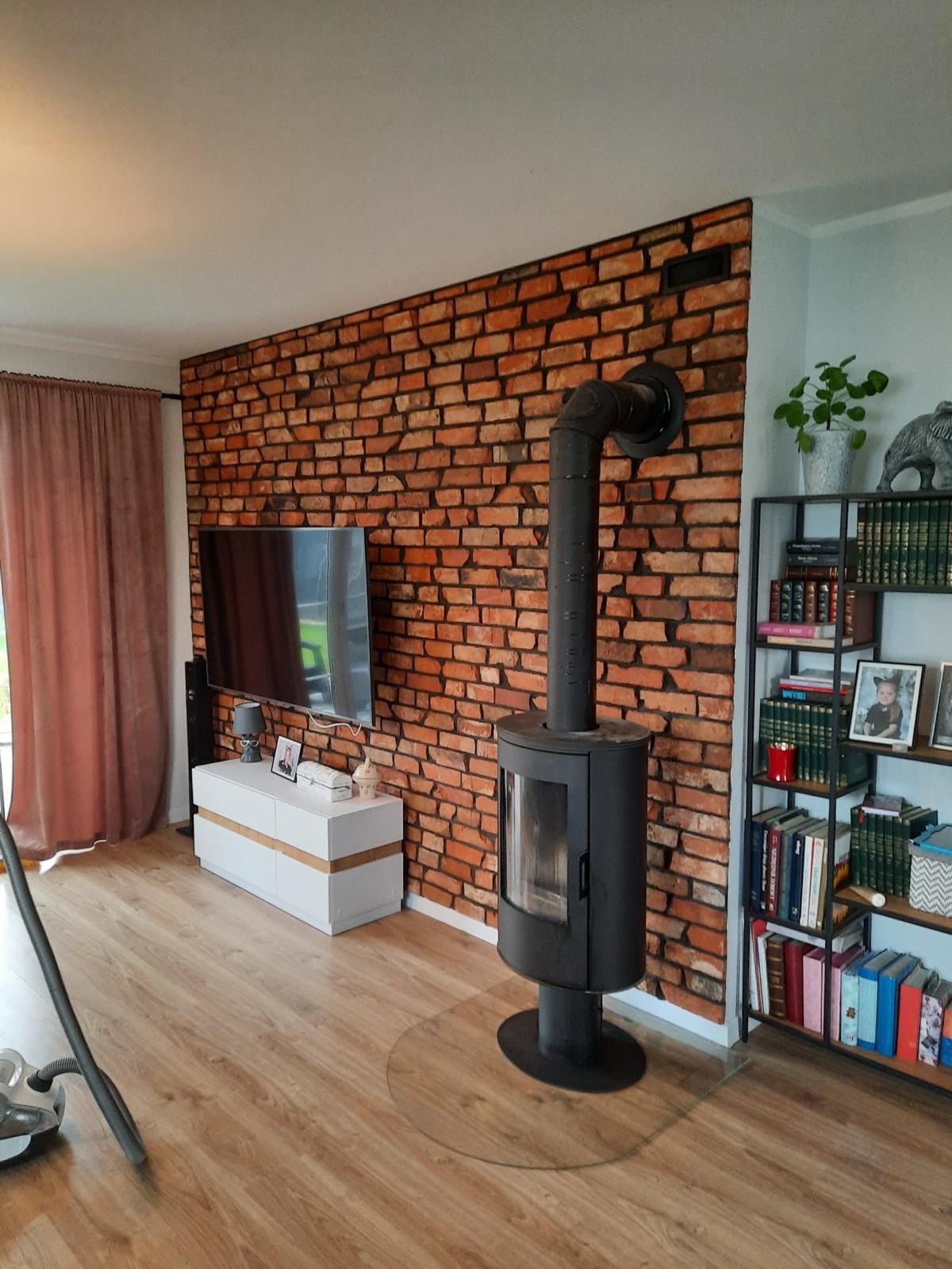 Salon z ceglaną ścianą, kominkiem wolnostojącym na szklanej podstawie, białą komodą pod telewizorem i regałem z książkami