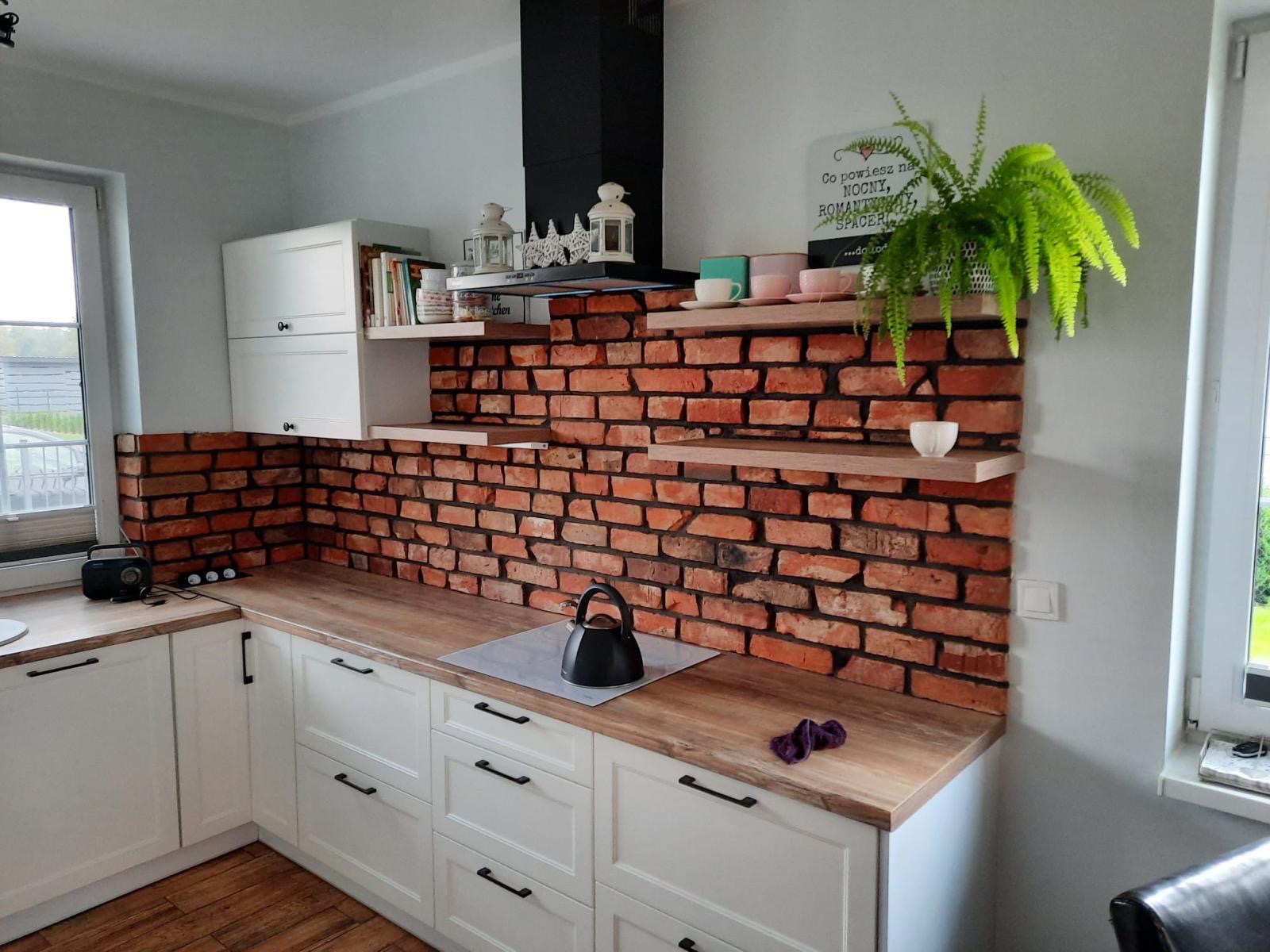 Kuchnia z białymi szafkami i blatem imitującym drewno, ściana wyłożona tapetą z motywem czerwonej cegły, na półkach dekoracje i roślina doniczkowa.