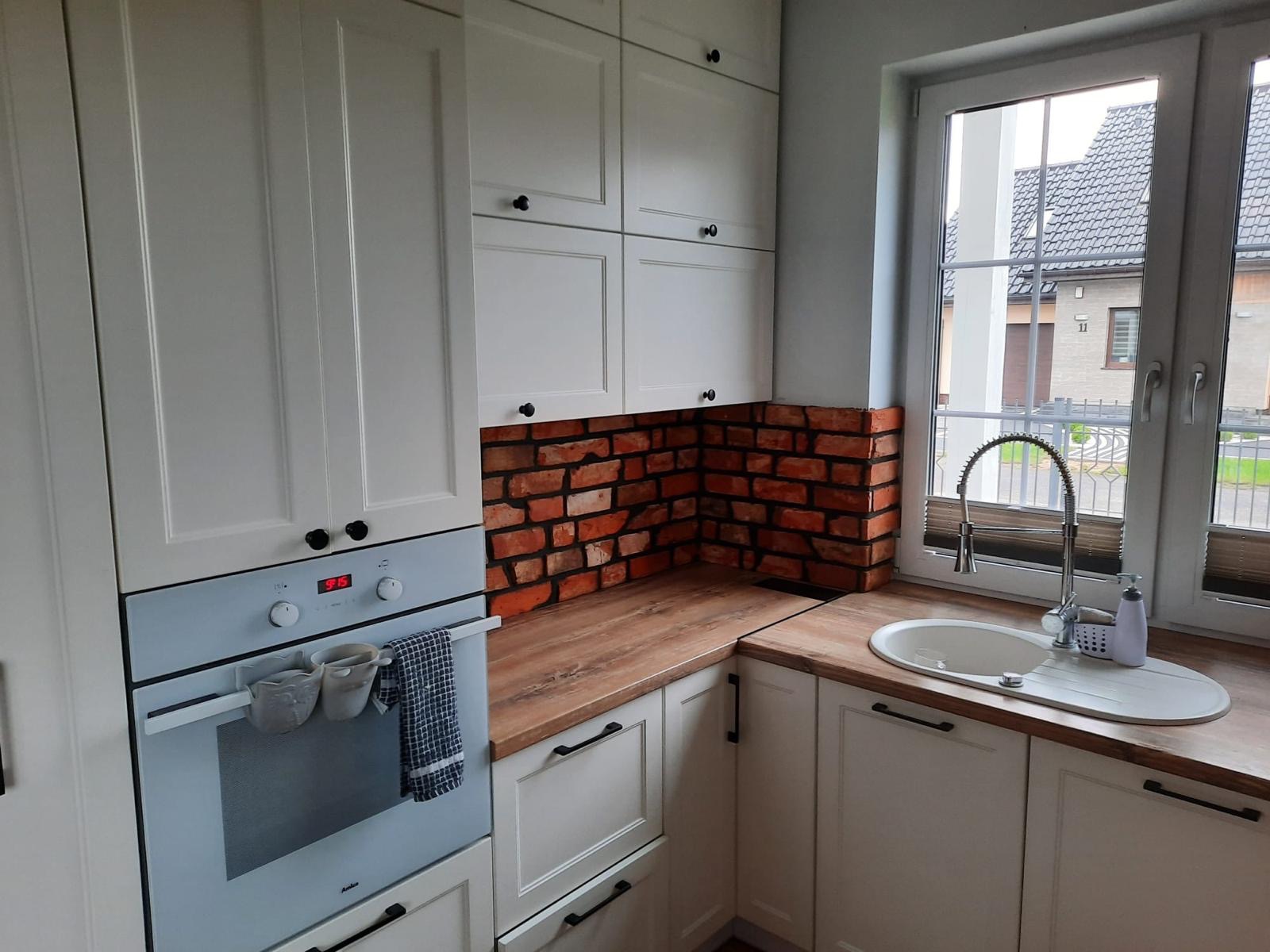 Kuchnia w stylu rustykalnym z białymi szafkami i ceglaną ścianą nad blatem, widok na zewnątrz przez okno z roletami.