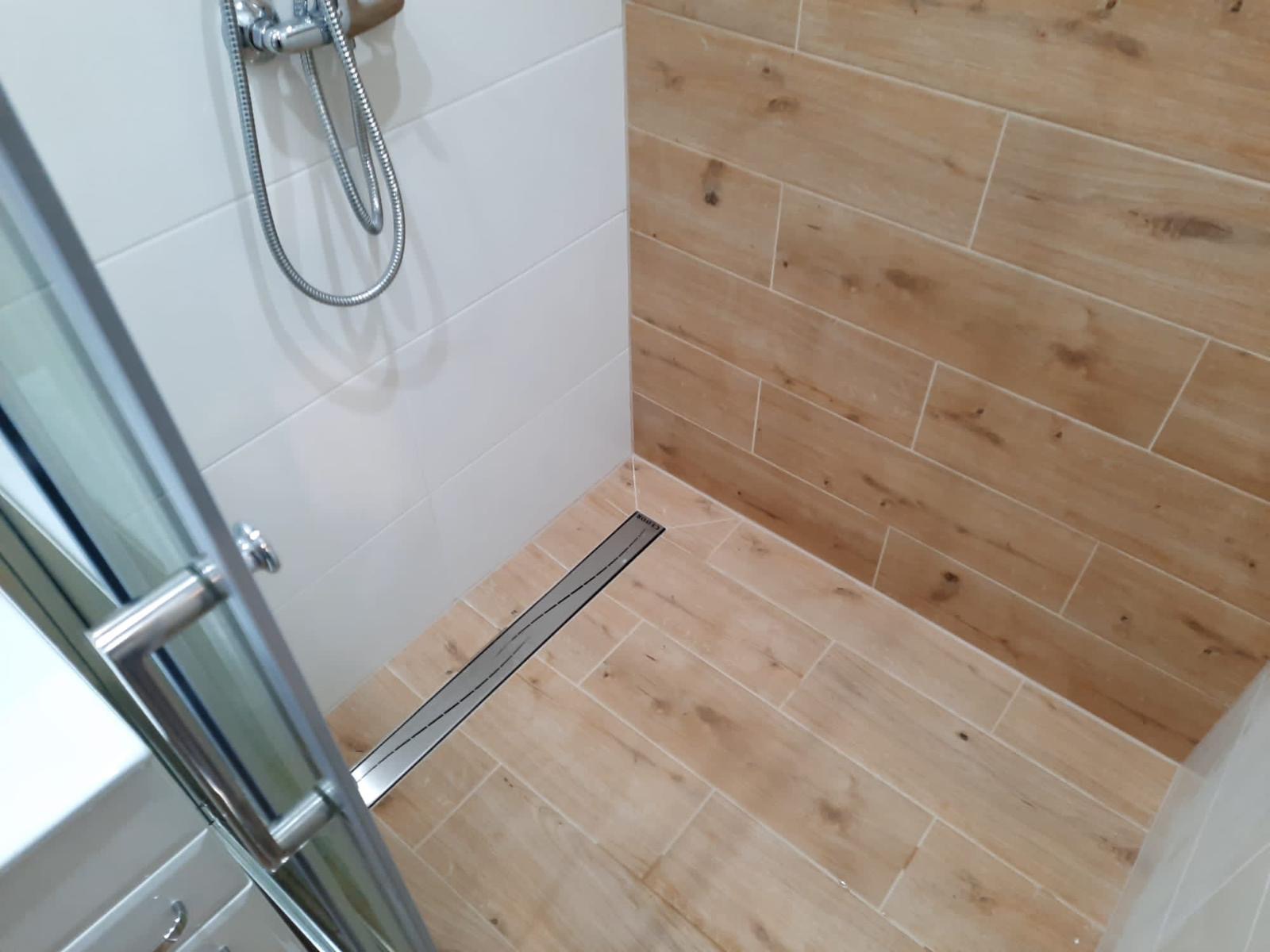 Nowoczesny prysznic z odpływem liniowym i płytkami imitującymi drewno na ścianie i podłodze, widoczna chromowana bateria i szklane drzwi.