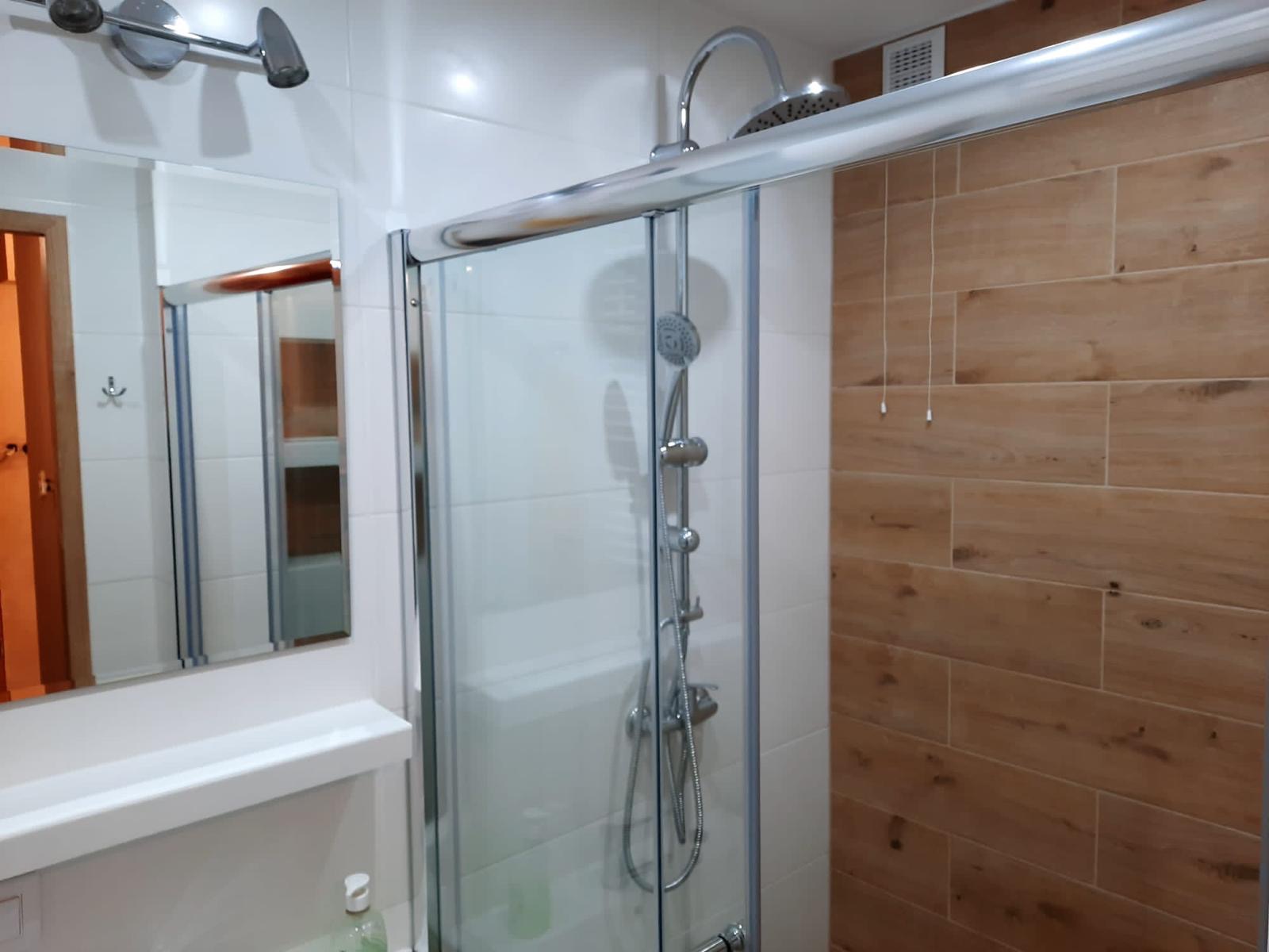 Kabina prysznicowa z przesuwnymi drzwiami, chromowanymi okuciami i deszczownicą, zamontowana w łazience z białymi płytkami i akcentem ściany z płytek imitujących drewno.