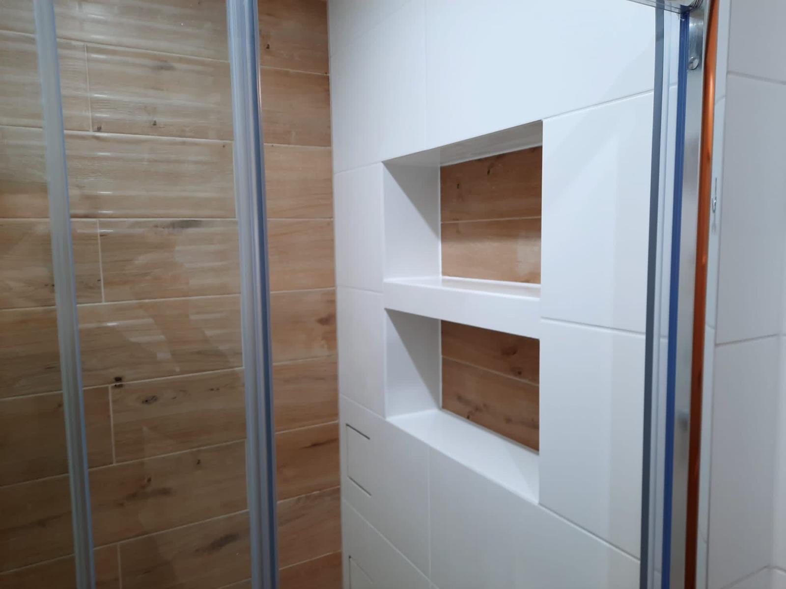 Wnęki w ścianie wyłożonej białymi płytkami, z drewnopodobnymi płytkami wewnątrz, widok przez szklane drzwi kabiny prysznicowej.