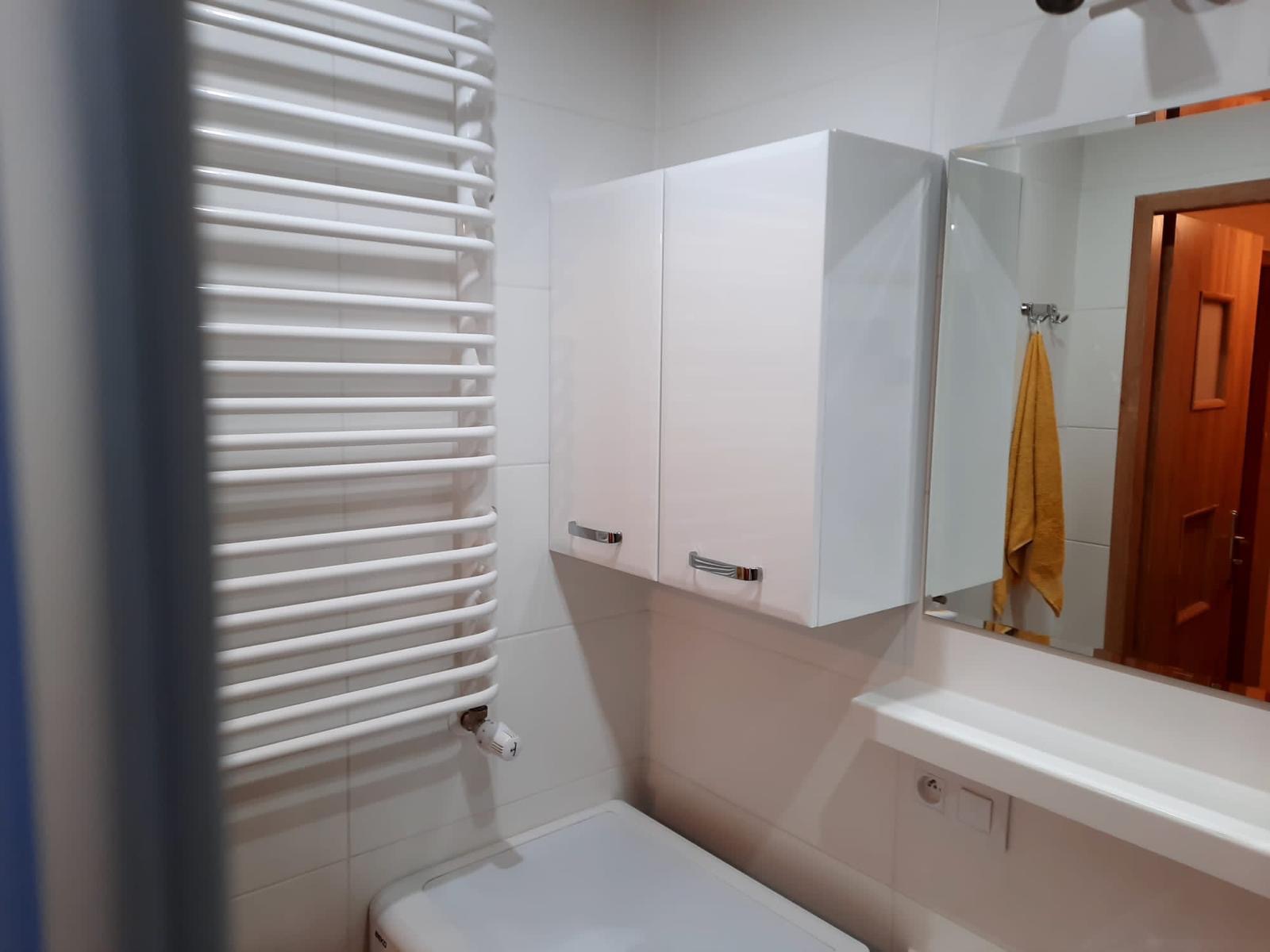 Biała łazienka z grzejnikiem drabinkowym, szafką wiszącą i lustrem, w którym odbija się drewniane drzwi.