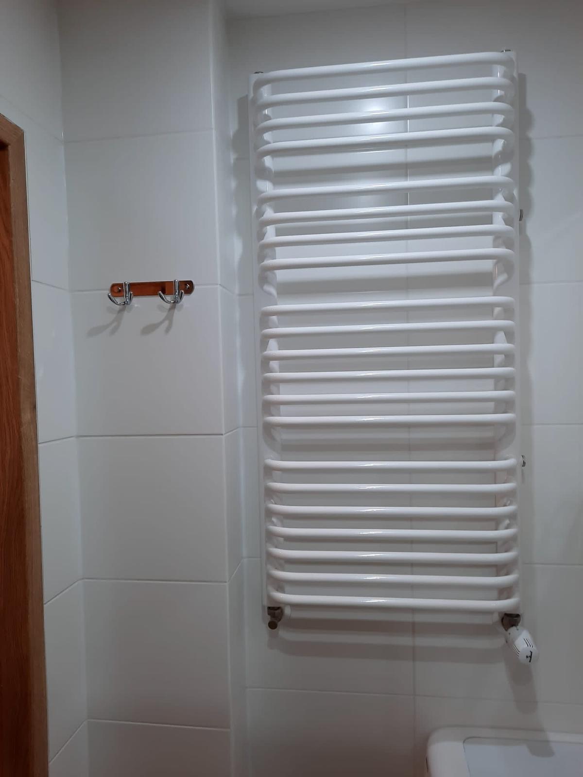 Biały grzejnik drabinkowy na ścianie z białych płytek w łazience, obok drzwi z drewnianą ościeżnicą i wieszak z dwoma haczykami.