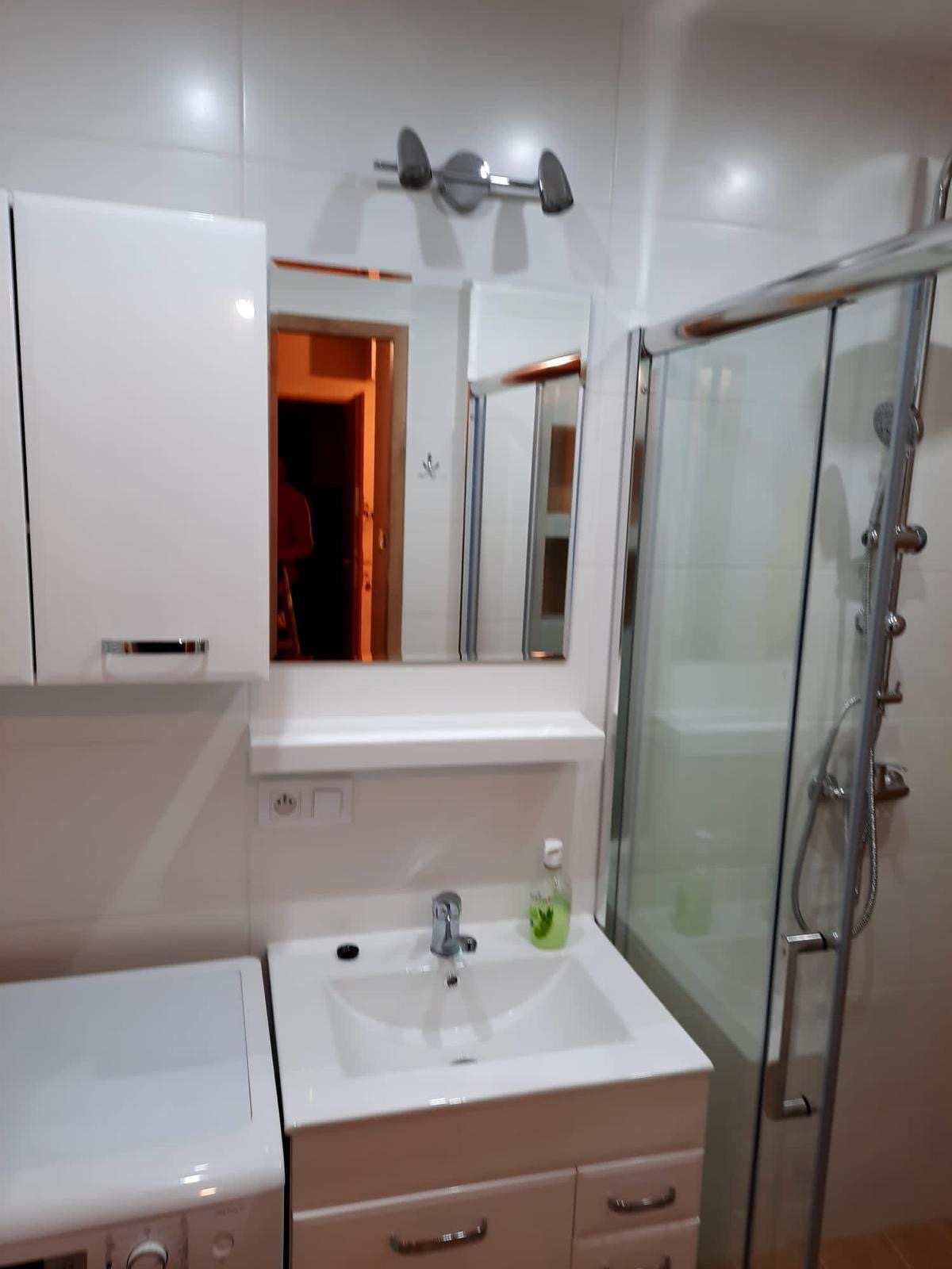 Jasna łazienka z kabiną prysznicową, umywalką z szafką, lustrem, szafką wiszącą i pralką, widoczne gniazdka elektryczne nad umywalką.