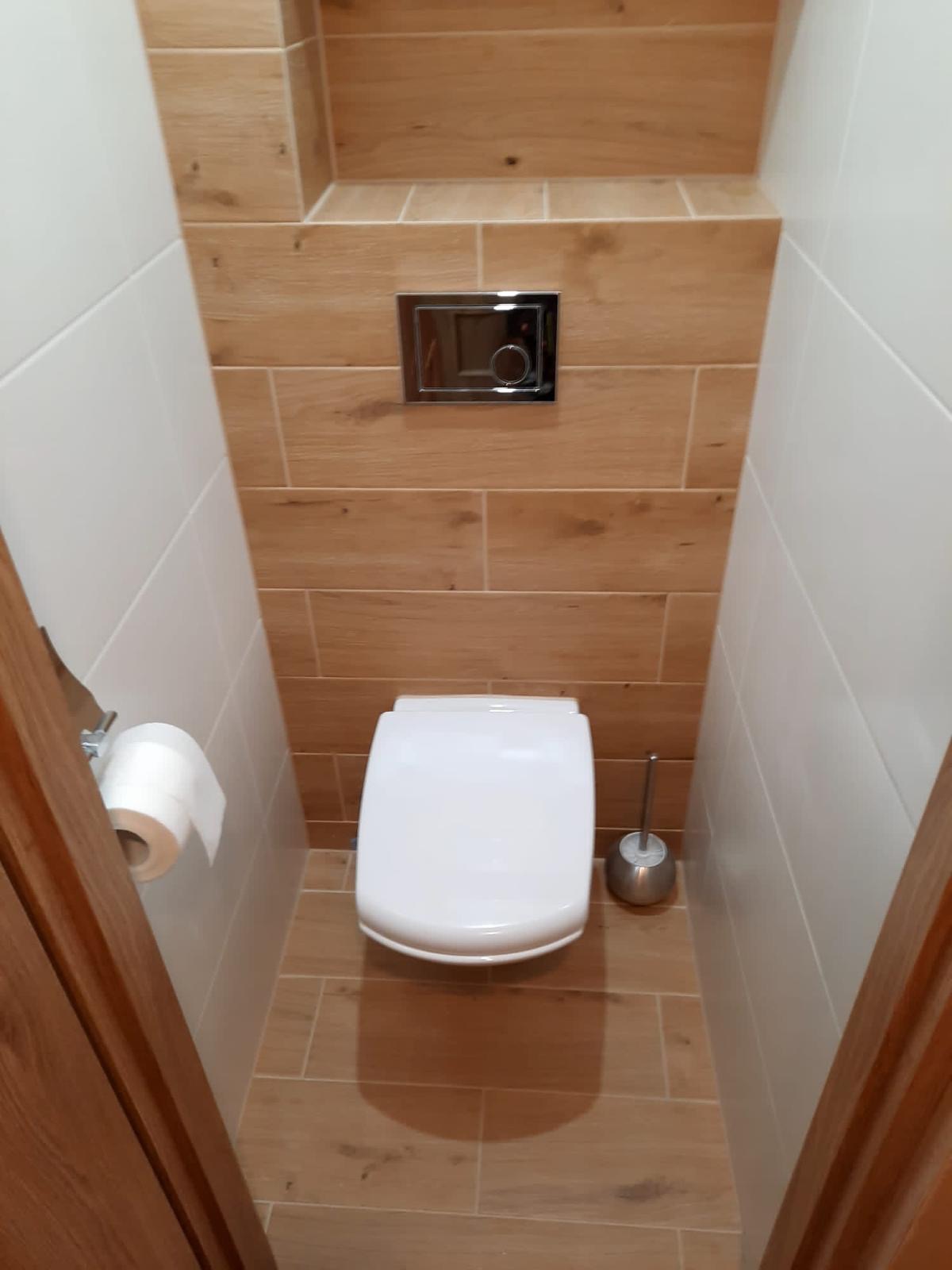 Wąska toaleta z białym kompaktem WC, chromowanym przyciskiem spłukiwania i drewnopodobnymi płytkami na ścianie akcentowej oraz białymi płytkami po bokach.
