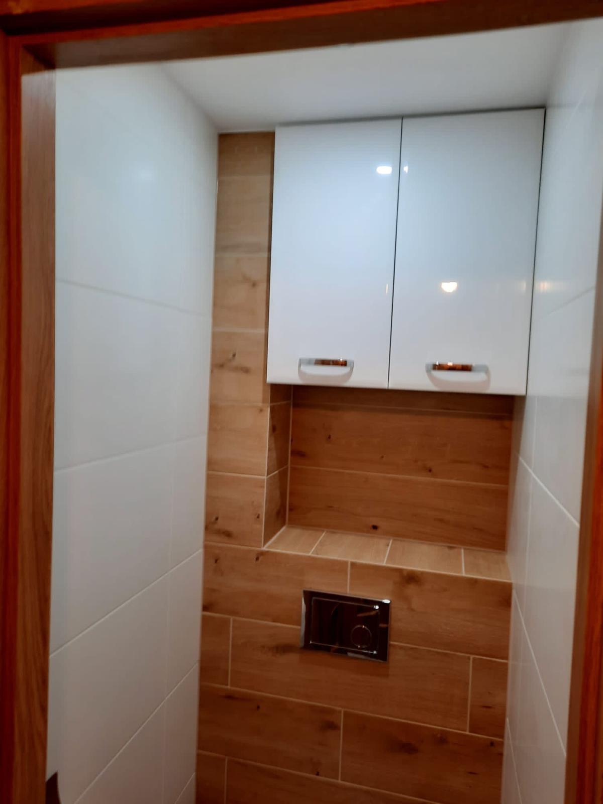 Nowoczesna toaleta z białymi płytkami i drewnopodobnymi panelami, wisząca szafka z białymi frontami i chromowanymi uchwytami nad zabudowaną spłuczką.