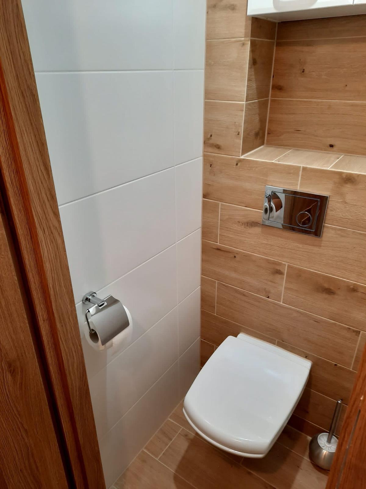 Nowoczesna toaleta z białymi i drewnopodobnymi płytkami, chromowanym uchwytem na papier toaletowy i przyciskiem spłukującym w ramce.