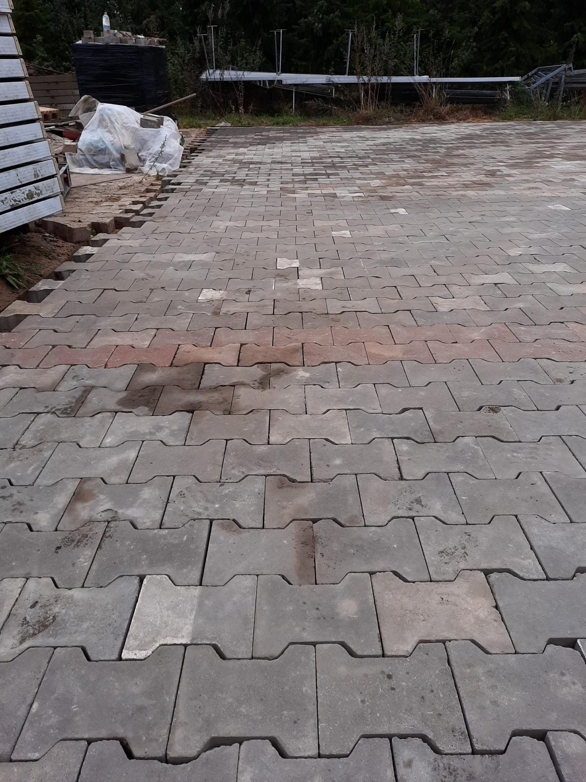 Układanie szarej kostki brukowej typu 'H' na podwórku, widoczne przygotowane podłoże i materiały budowlane w tle.