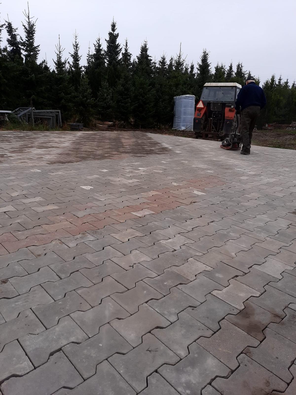 Układanie kostki brukowej na posesji, widoczny pracownik z zagęszczarką i traktor Zetor w tle, obok leżą spakowane kostki