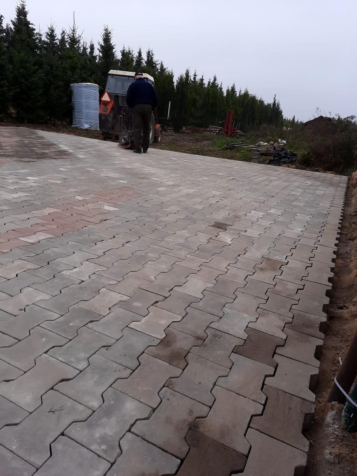 Układanie szarej kostki brukowej typu I na posesji, widoczny ciągnik rolniczy z tyłu i przygotowane podłoże po prawej stronie.