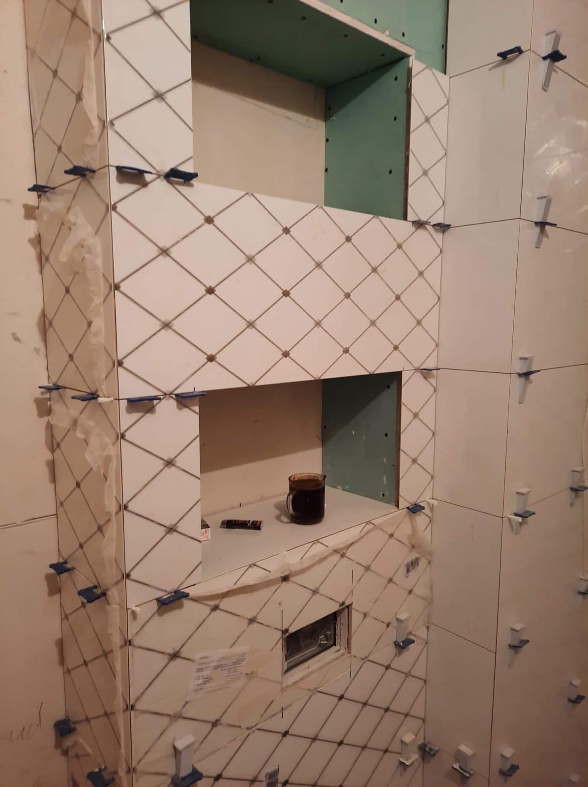 Układanie białych płytek z romboidalnym wzorem w łazience, widoczne niebieskie krzyżyki dystansowe, kubek z kawą i długopis na półce wnęki.