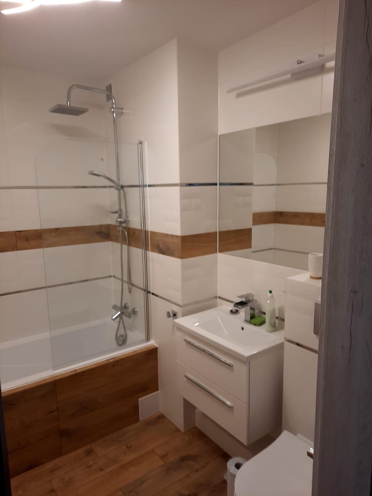 Nowoczesna łazienka z białymi płytkami imitującymi cegłę, drewnianymi akcentami i kabiną prysznicową z szybą oraz białą szafką pod umywalką i lustrem.