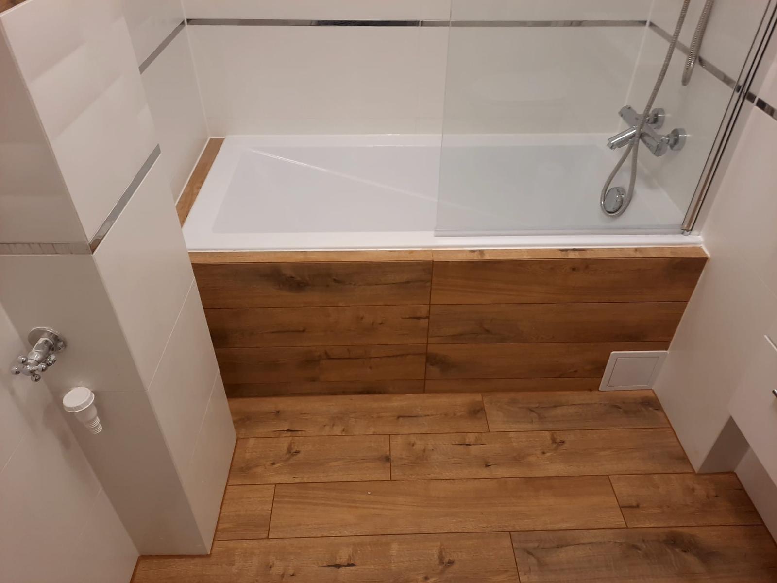 Nowoczesna łazienka z drewnopodobną okładziną wanny i na podłodze, białe płytki ścienne z metalowymi listwami dekoracyjnymi, szklany parawan nawannowy.