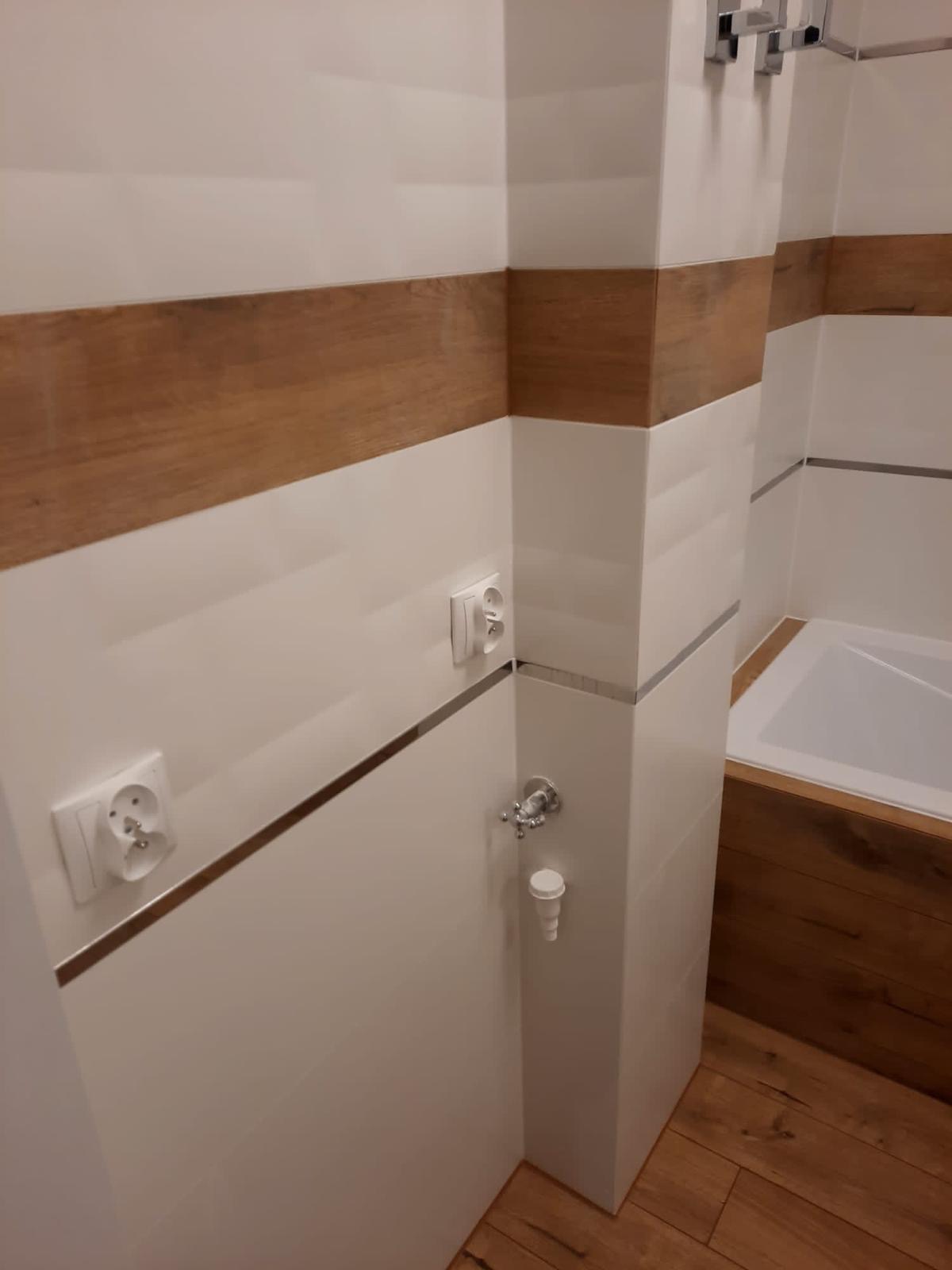 Wykończona łazienka z białymi płytkami i poziomym pasem dekoracyjnym imitującym drewno, widoczne gniazdka elektryczne i zawór.