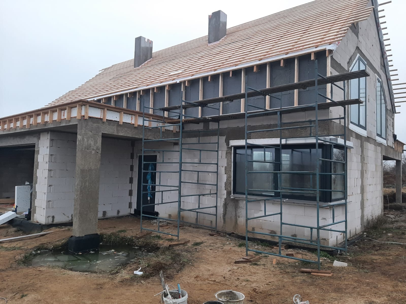 Wznoszony dom murowany z białych bloczków, widoczne rusztowania, przygotowywane drewniane elementy dachu, czarne okna, teren budowy z kałużą.
