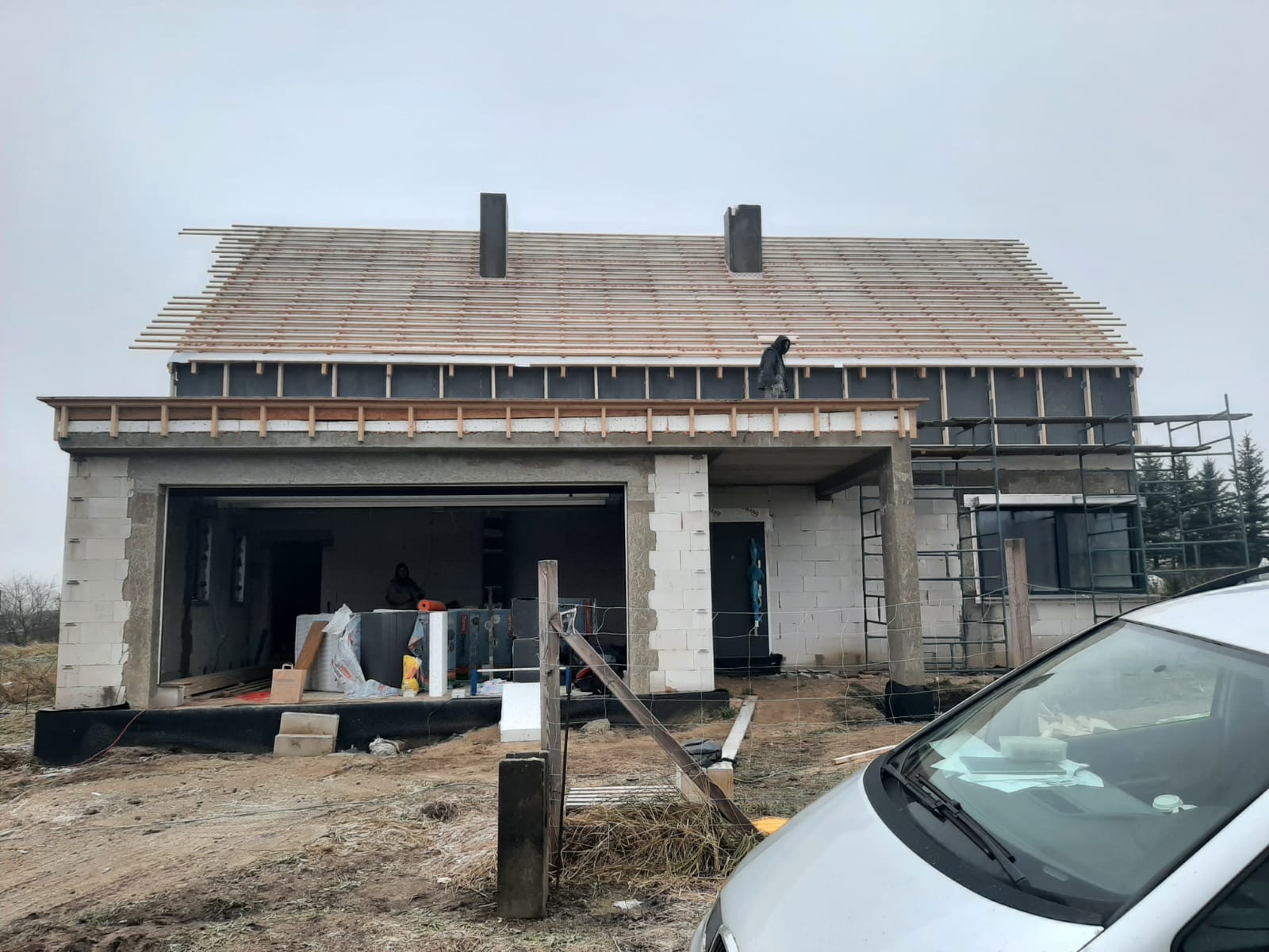 Surowy stan budynku z bloczków betonowych w trakcie budowy dachu, widoczne rusztowania, częściowo ułożone deski dachowe i materiały budowlane w garażu, na pierwszym planie fragment samochodu.