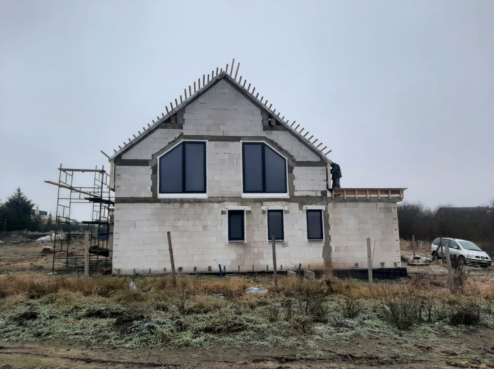 Surowy stan domu jednorodzinnego z bloczków, z zamontowanymi oknami, widoczne rusztowania i praca na dachu, pochmurne niebo w tle.