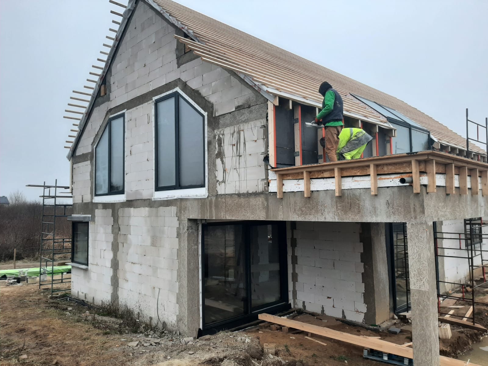 Budowa dwukondygnacyjnego domu z białych bloczków, widoczne duże okna z czarnymi ramami, drewniana konstrukcja dachu w trakcie montażu, robotnicy pracujący na drewnianym rusztowaniu przy montażu...