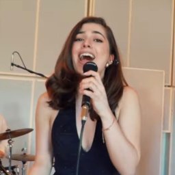 MUZYCZNY WARSZTAT PRZEMYSŁAW KUTA - Wokalistka w czarnej sukience śpiewa do mikrofonu na scenie. W tle widoczny perkusista i panele akustyczne. Jasne oświetlenie, dynamiczne ujęcie.