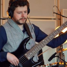 MUZYCZNY WARSZTAT PRZEMYSŁAW KUTA - Basista w kamizelce gra na czarnej gitarze basowej, w tle perkusja i panele akustyczne. Członek zespołu muzycznego podczas występu lub nagrania w studio.