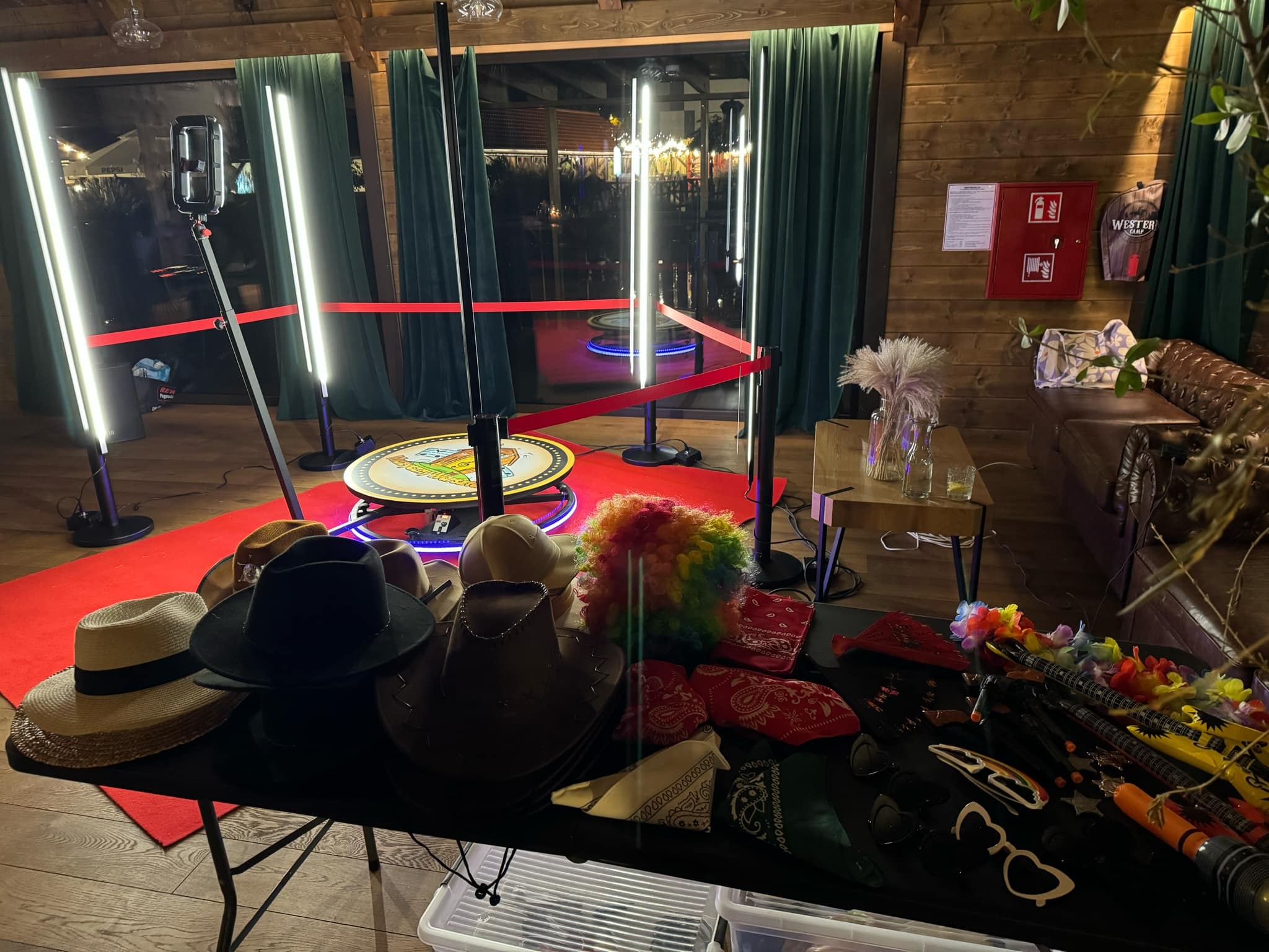 Stanowisko z fotobudką: rekwizyty (kapelusze, peruki, okulary, bandany), czerwony dywan, oświetlenie LED, drewniane wnętrze.