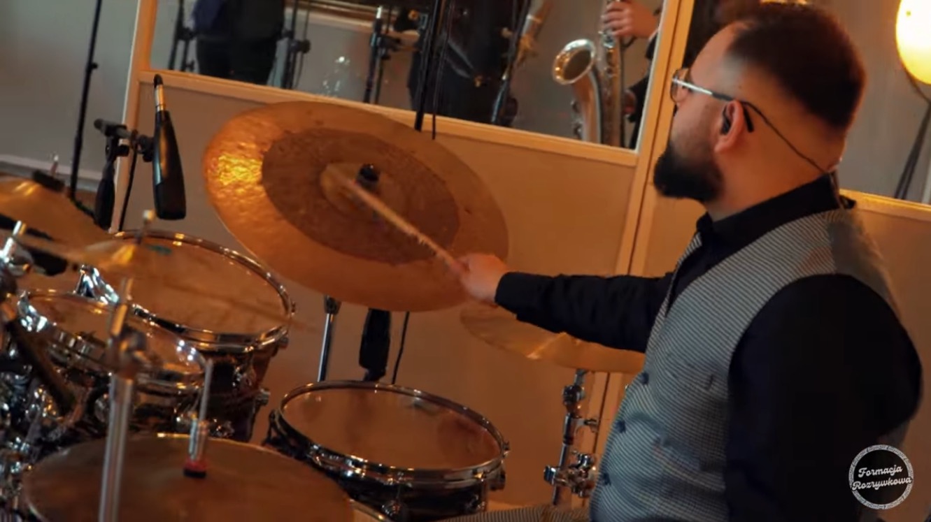 Perkusista w akcji uderza pałeczką w talerz perkusyjny. W tle widoczny saksofonista. Profesjonalne nagłośnienie i oświetlenie sceniczne.