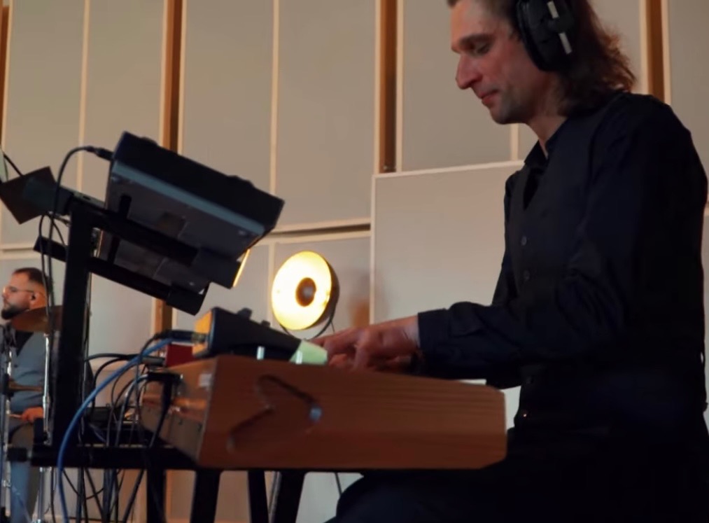 Muzyk w czarnej kamizelce gra na klawiszach w studiu nagraniowym, widoczny perkusista w tle. Nietypowe oświetlenie i drewniana obudowa klawiszy.