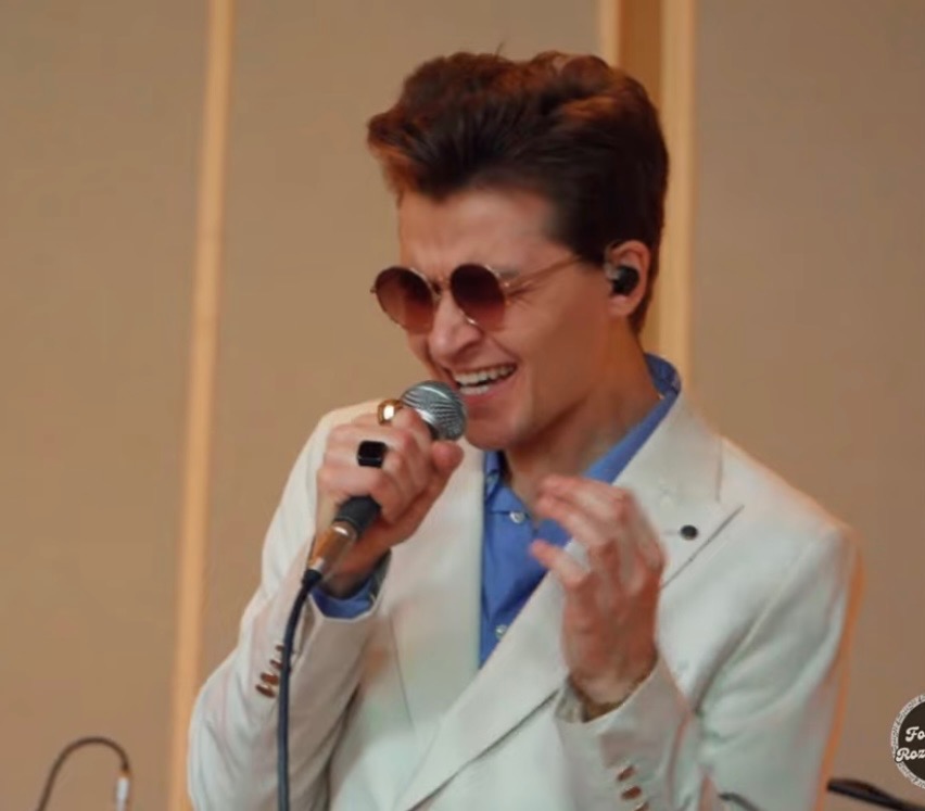 Uśmiechnięty wokalista w okularach przeciwsłonecznych, z mikrofonem w dłoni, ubrany w biały garnitur, występuje na scenie. Widoczne czarne słuchawki w uszach.