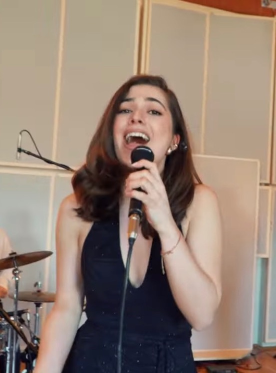 Wokalistka w czarnej sukience śpiewa do mikrofonu na scenie. W tle widoczny perkusista i panele akustyczne. Jasne oświetlenie, dynamiczne ujęcie.