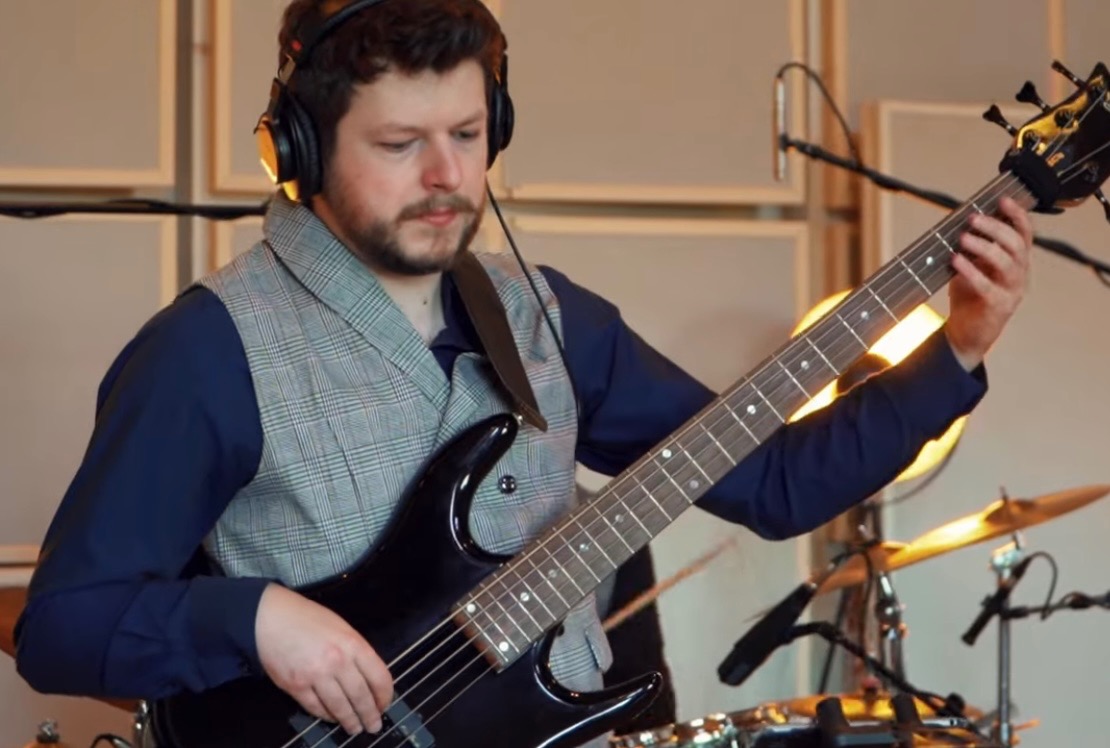 Basista w kamizelce gra na czarnej gitarze basowej, w tle perkusja i panele akustyczne. Członek zespołu muzycznego podczas występu lub nagrania w studio.