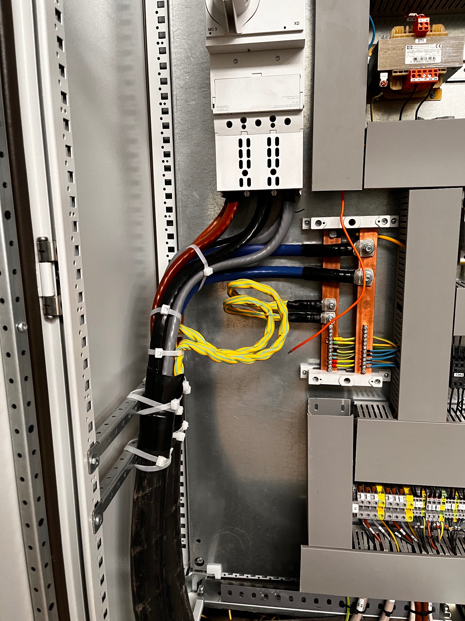 Wnętrze szafy elektrycznej z widocznymi przewodami w różnych kolorach, zabezpieczonymi opaskami zaciskowymi, oraz elementami sterowniczymi i transformatorem.