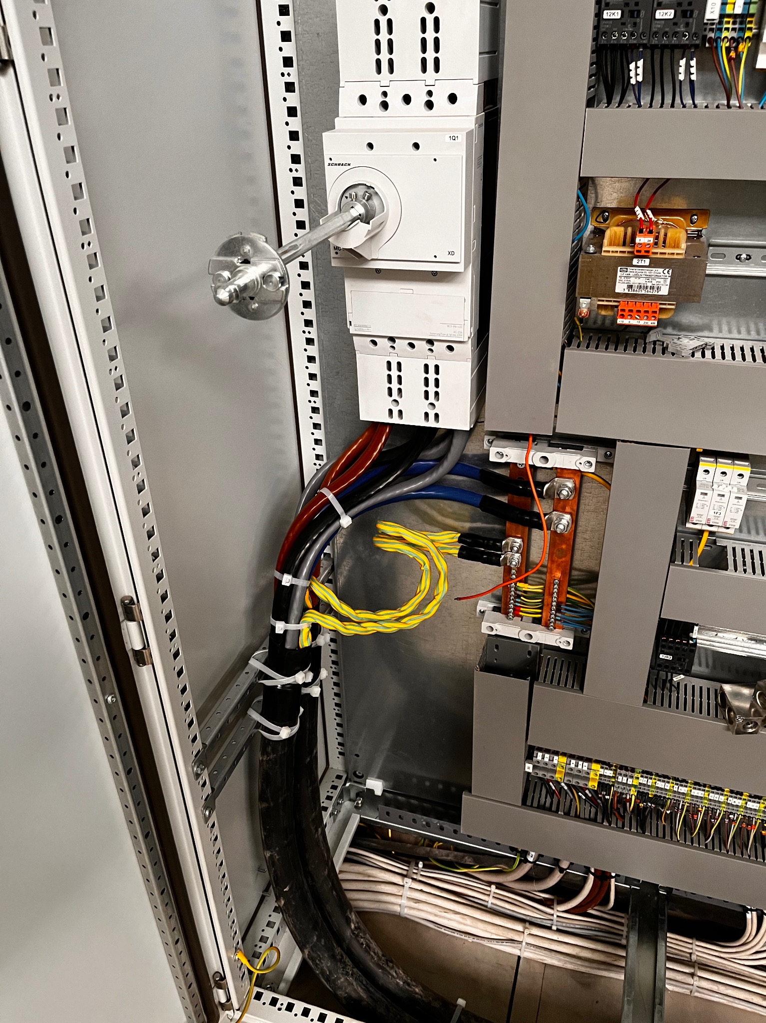 Wnętrze szafy sterowniczej z widocznymi elementami: wyłącznik główny z dźwignią, transformator, listwy zaciskowe i uporządkowane przewody w różnych kolorach, zabezpieczone opaskami kablowymi.