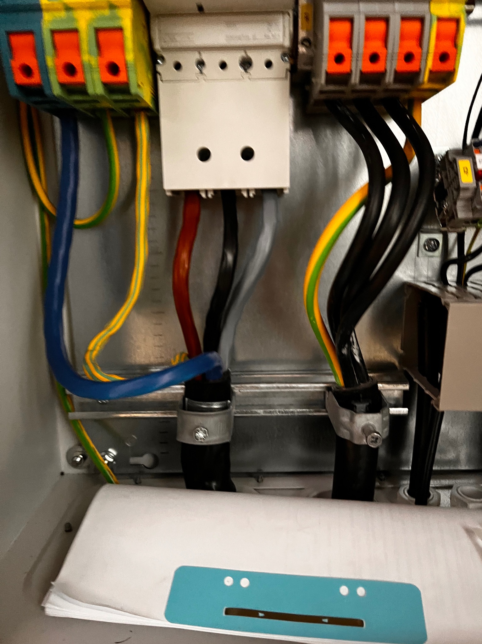 Widok z bliska wnętrza skrzynki rozdzielczej z kolorowymi przewodami podłączonymi do zacisków i zabezpieczonymi uchwytami, widoczne dokumenty na dnie skrzynki.