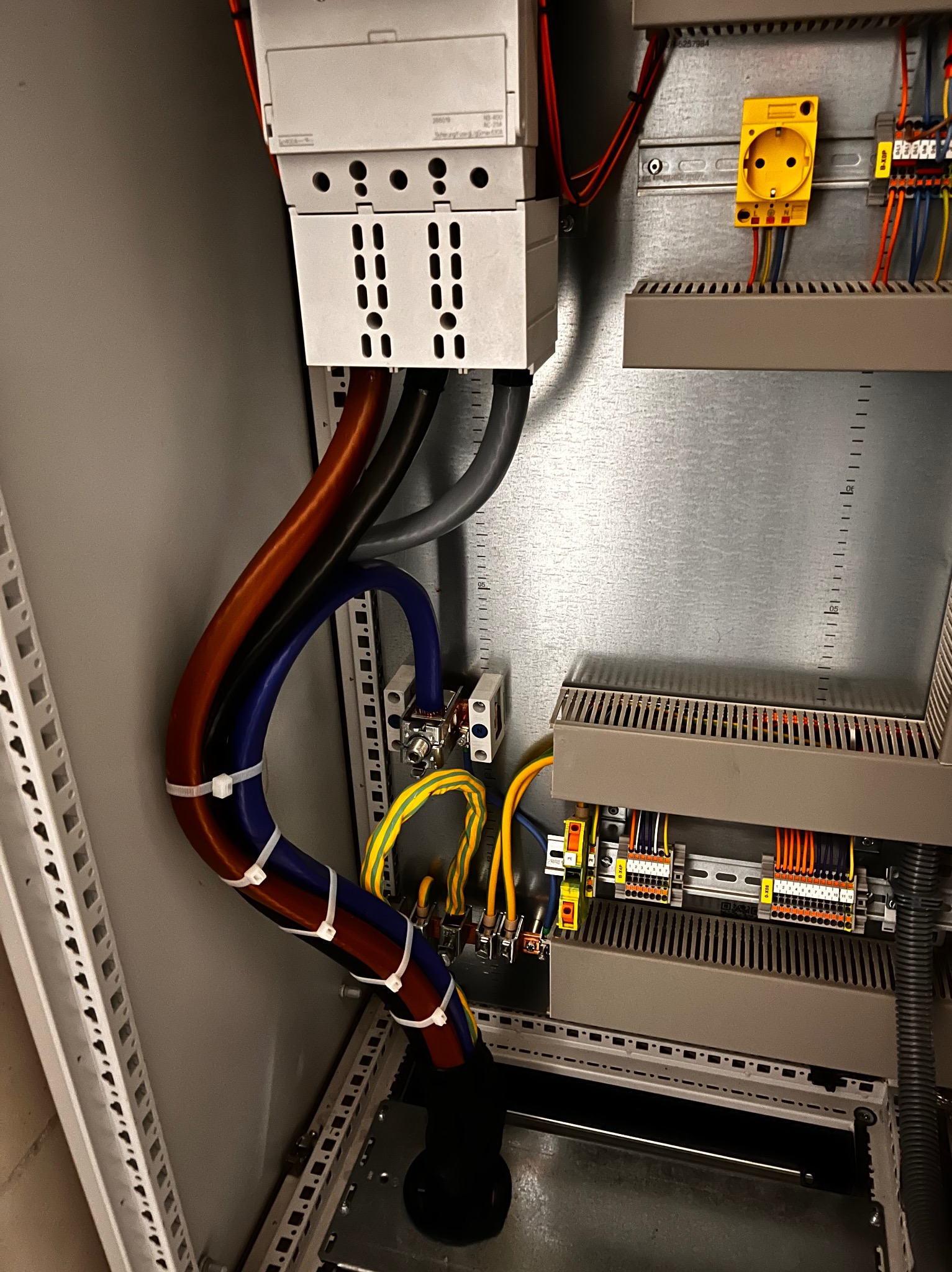 Wnętrze szafy elektrycznej z widocznymi przewodami o różnych kolorach (brązowy, niebieski, czarny, żółto-zielony) spiętymi białymi opaskami, modułami DIN, gniazdem elektrycznym i listwami zaciskowymi.