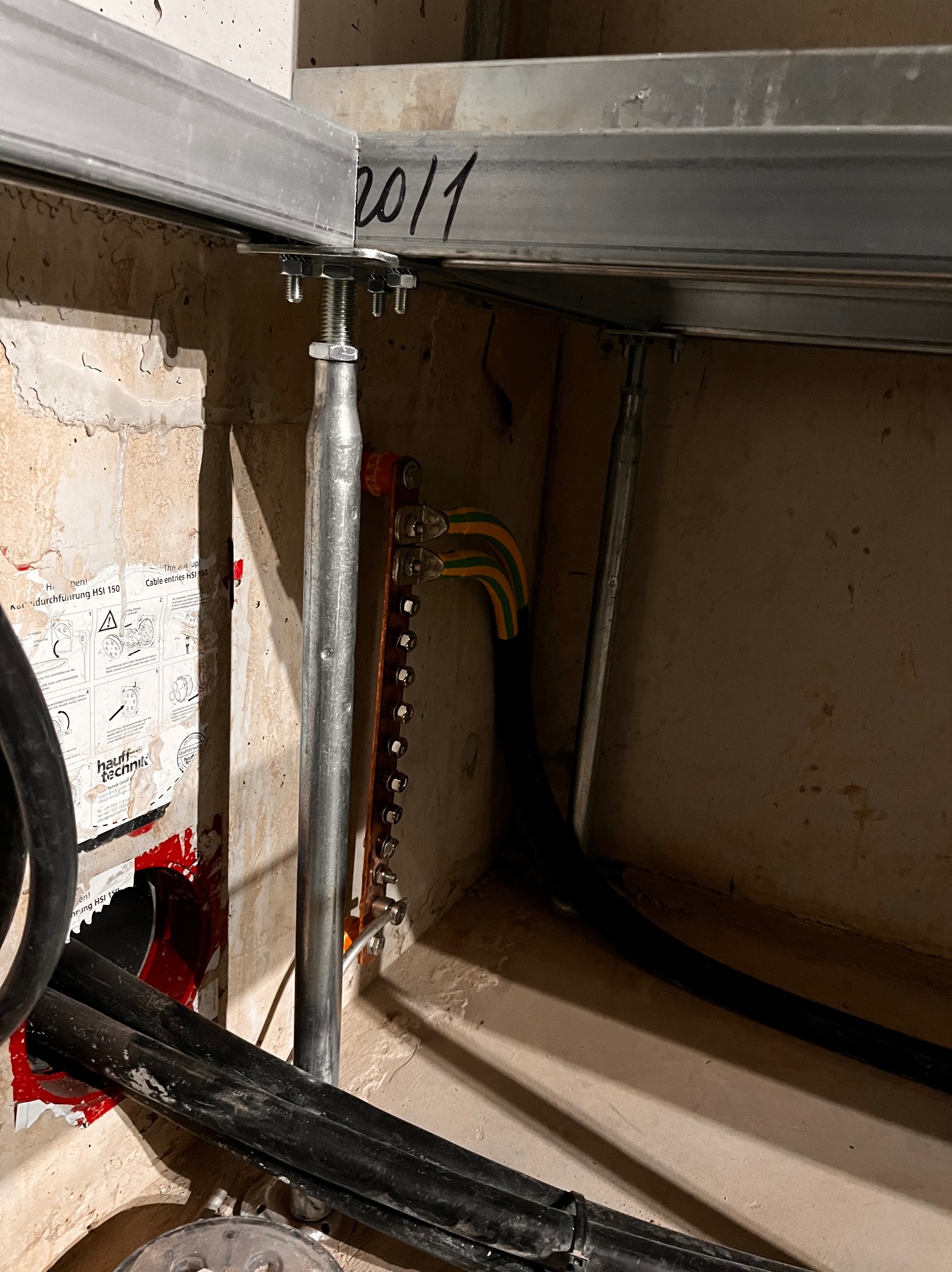 Metalowa szyna montażowa przymocowana do sufitu, z widocznymi przewodami i listwą zaciskową uziemiającą, w tle ściana z naklejką informacyjną producenta okablowania.