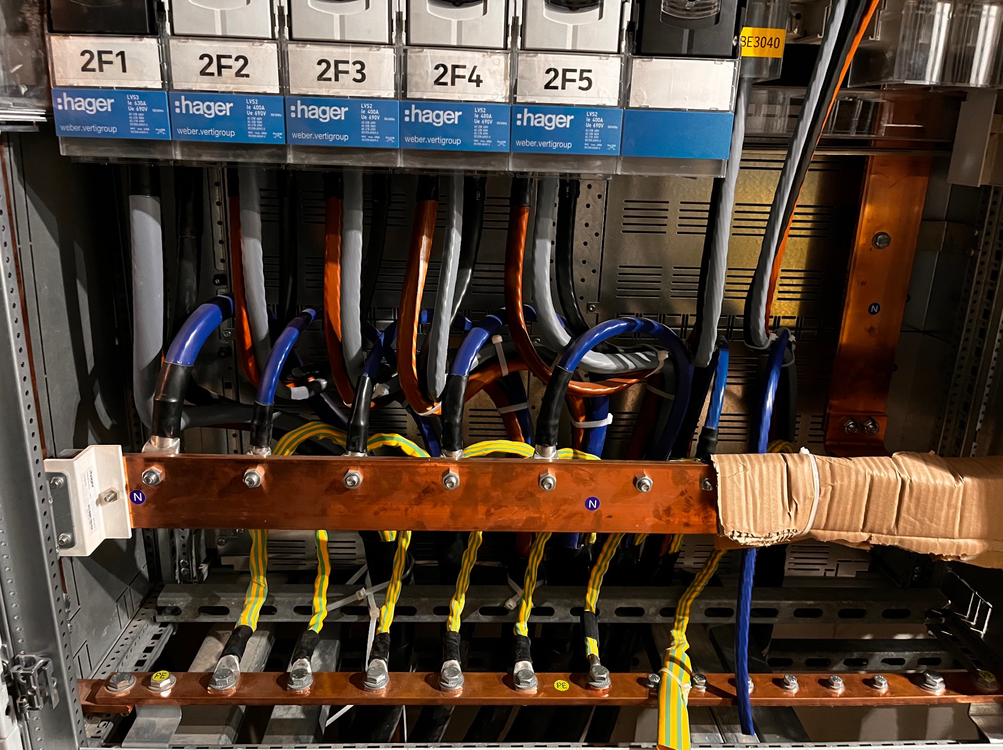 Rozdzielnica elektryczna z widocznymi bezpiecznikami Hager, miedzianymi szynami zbiorczymi i przewodami w różnych kolorach, w tym niebieskim, szarym, pomarańczowym i żółto-zielonym, z oznaczeniami...