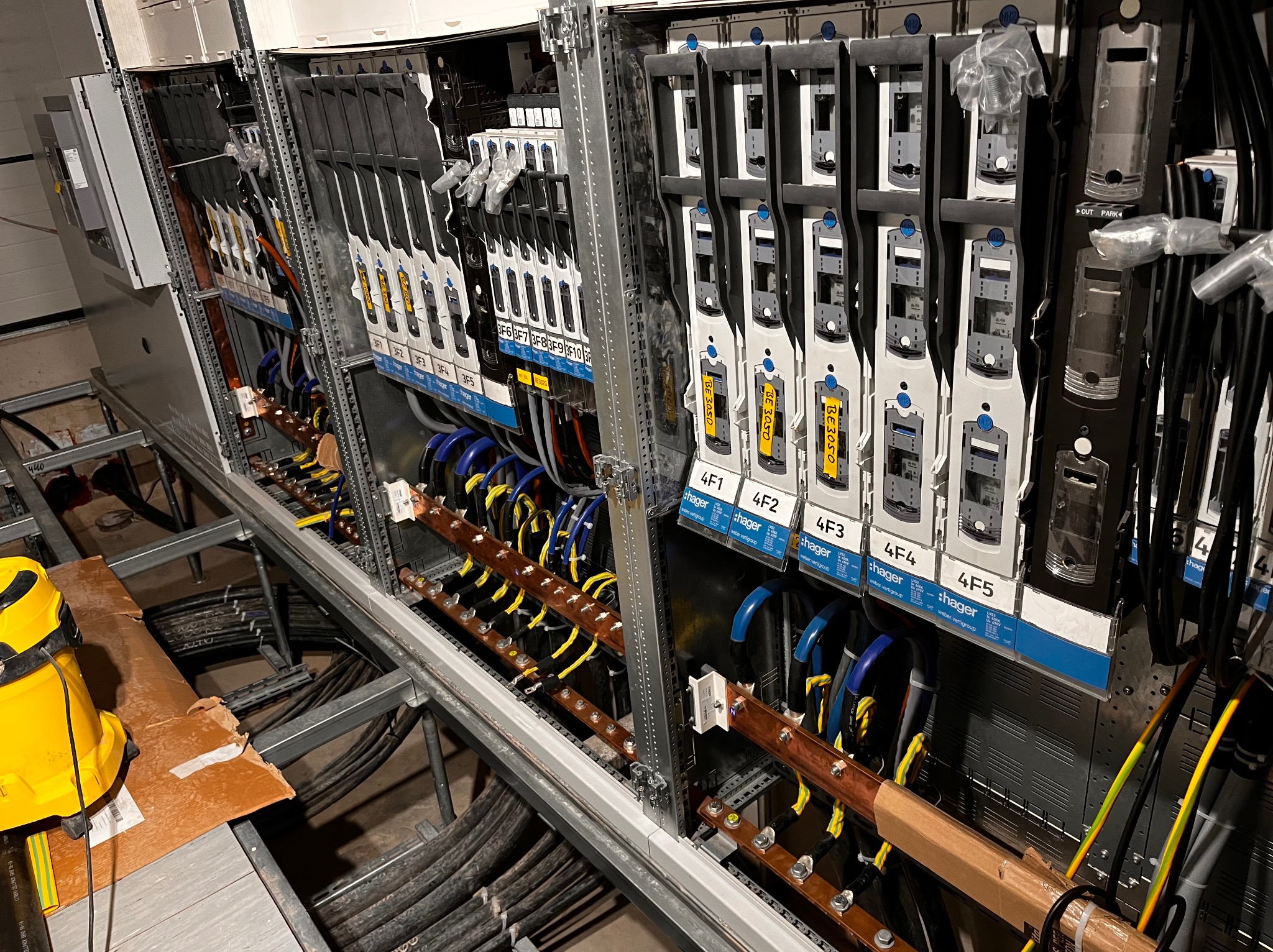 Rozdzielnia elektryczna w trakcie montażu, widoczne szyny zbiorcze miedziane, zabezpieczenia modułowe marki Hager, okablowanie w kolorach niebieskim i żółto-zielonym, żółty odkurzacz przemysłowy...