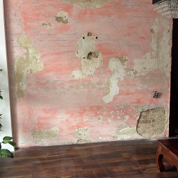 Kamil Kochanowski - Ściana w trakcie przygotowania do malowania: stara farba odpryskuje, ukazując warstwy poprzednich kolorów i nierówności podłoża, widoczne gniazdko elektryczne.