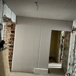 Kamil Kochanowski - Wnętrze w trakcie remontu: ściany z płyt gipsowo-kartonowych w surowym stanie, odsłonięta cegła, widoczne pozostałości starej tapety, sufit z wystającymi przewodami, gruz na podłodze, drabina...