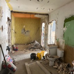 Kamil Kochanowski - Wnętrze mieszkania w trakcie generalnego remontu, widoczne ślady po zerwanych tapetach, resztki gruzu, odsłonięte instalacje sufitowe, częściowo zdemontowana toaleta i umywalka, worki z zaprawą...