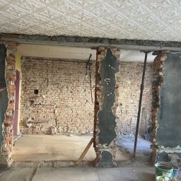 Kamil Kochanowski - Zdjęcie przedstawia wnętrze w trakcie demontażu ścian działowych z odsłoniętą cegłą, widoczne podparte stalowe belki stropowe, fragment dekoracyjnego sufitu kasetonowego, oraz narzędzia pracy...