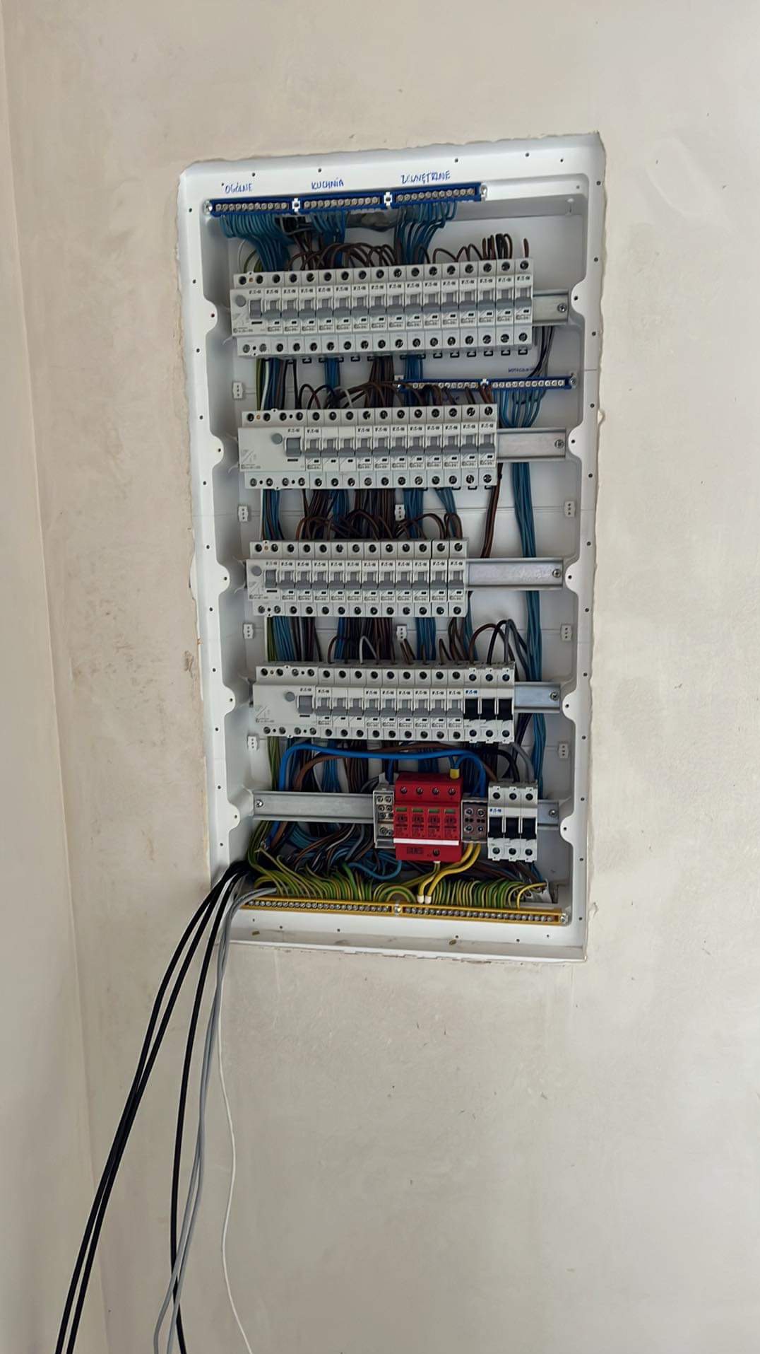 Rozdzielnica elektryczna wnękowa z zainstalowanymi bezpiecznikami, przewodami o różnych kolorach i oznaczeniami 'ogólne', 'kuchnia', 'zewnętrzne' na górze, zamontowana na surowej ścianie.