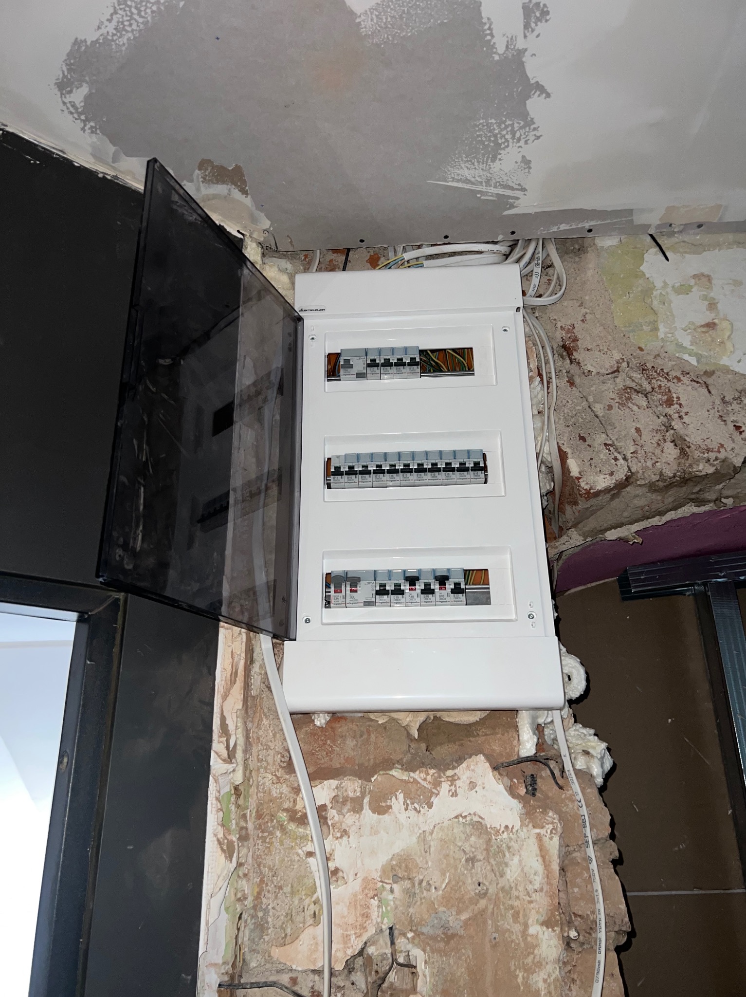 Nowa, biała skrzynka z bezpiecznikami zamontowana na surowej ścianie z widocznymi przewodami i pianką montażową, z otwartą, przyciemnioną klapką.