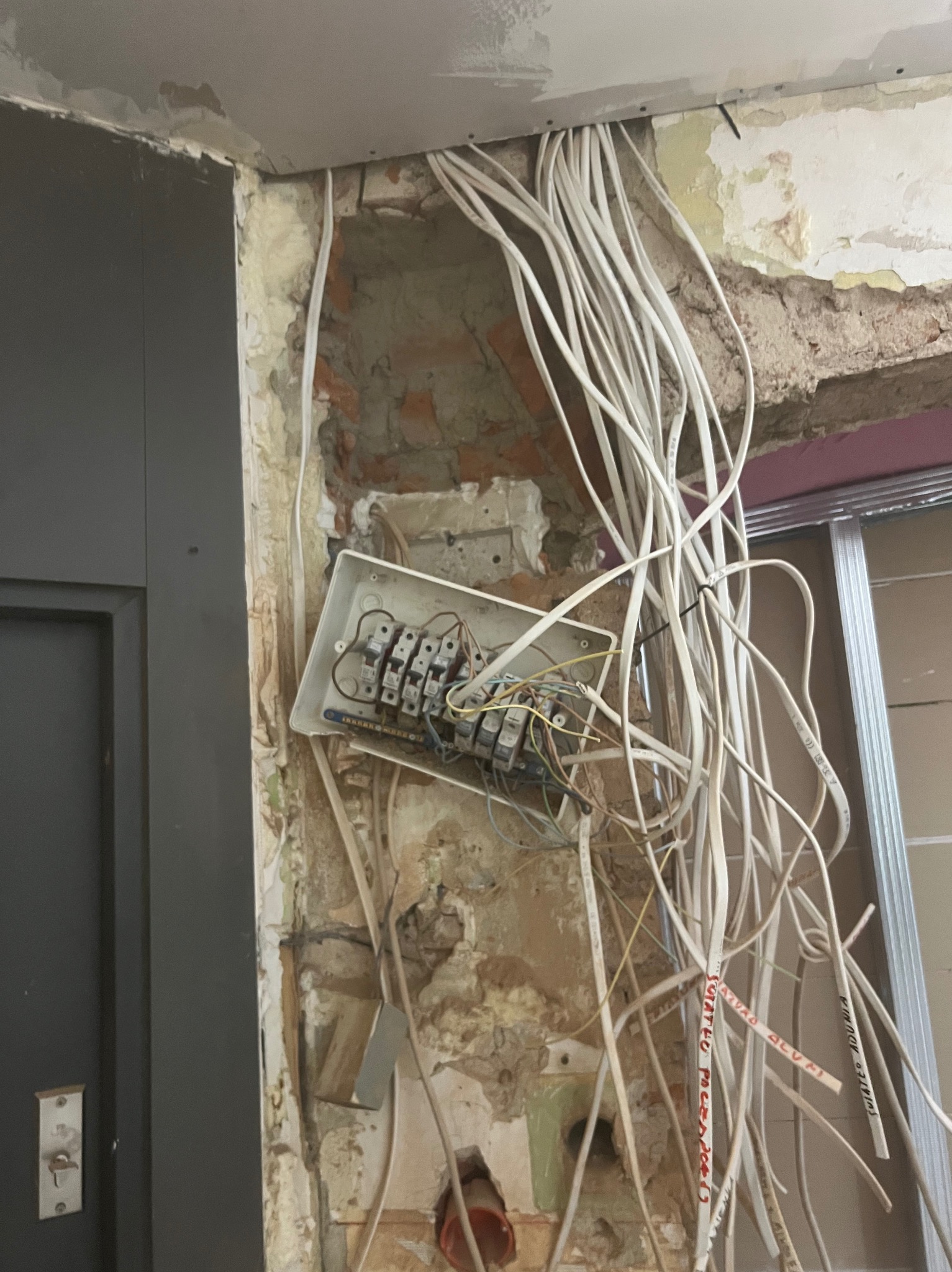 Otwarta skrzynka z bezpiecznikami i wiązka kabli wystających ze ściany podczas remontu, widoczne fragmenty cegieł i resztki tynku.