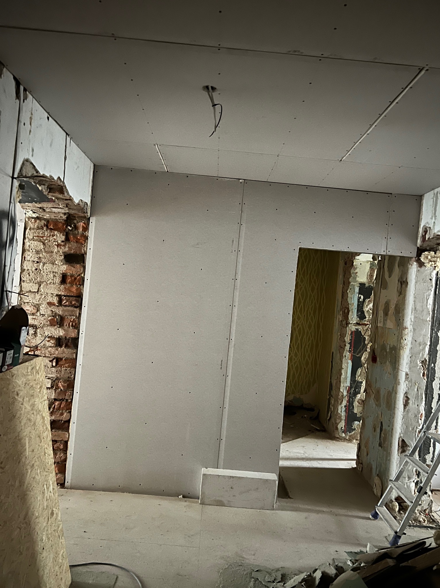 Wnętrze w trakcie remontu: ściany z płyt gipsowo-kartonowych w surowym stanie, odsłonięta cegła, widoczne pozostałości starej tapety, sufit z wystającymi przewodami, gruz na podłodze, drabina...